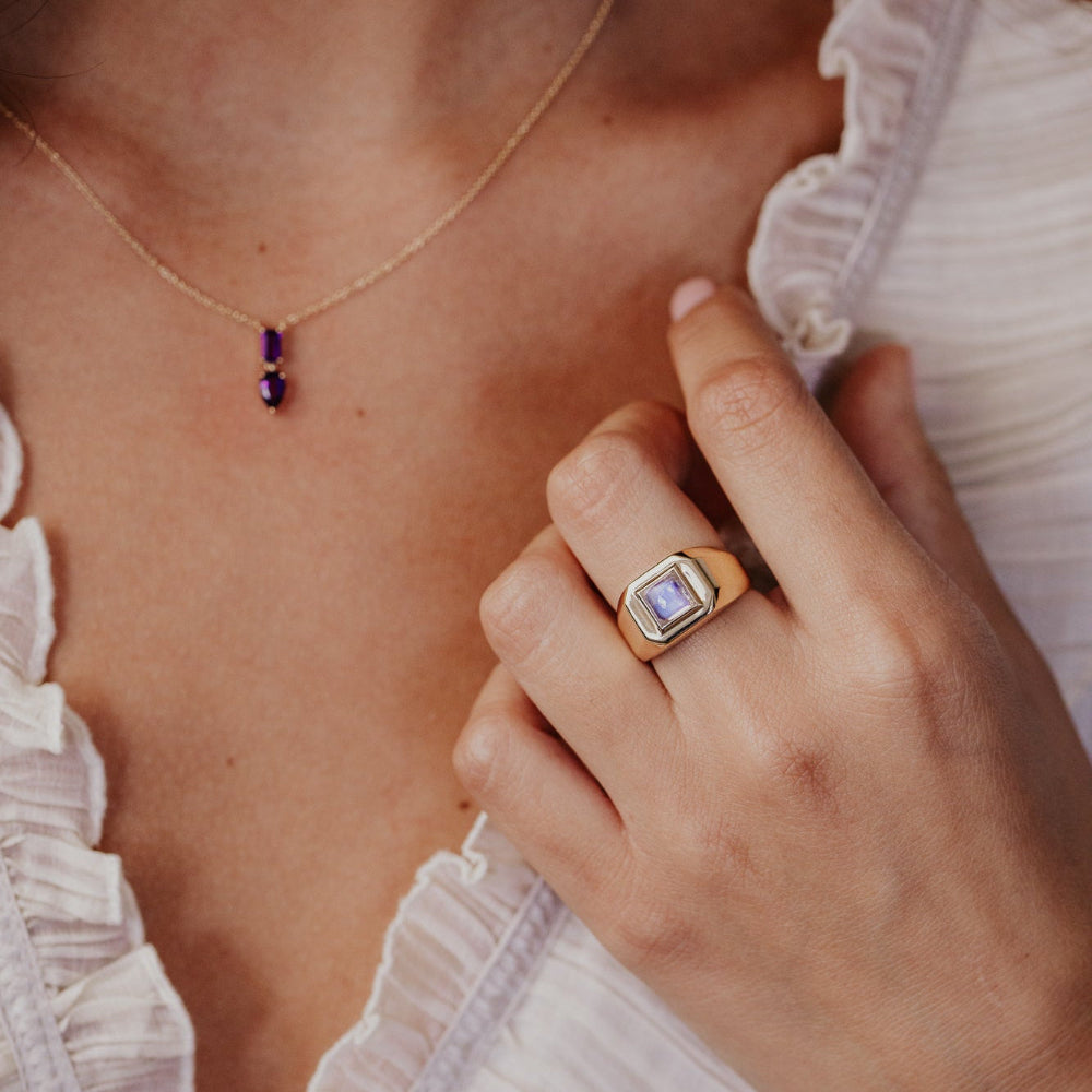 Close-up of a hand wearing a gold ring with a purple gemstone, with a blurred background.