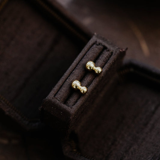 Gold double ball shaped earrings on a dark brown fabric background