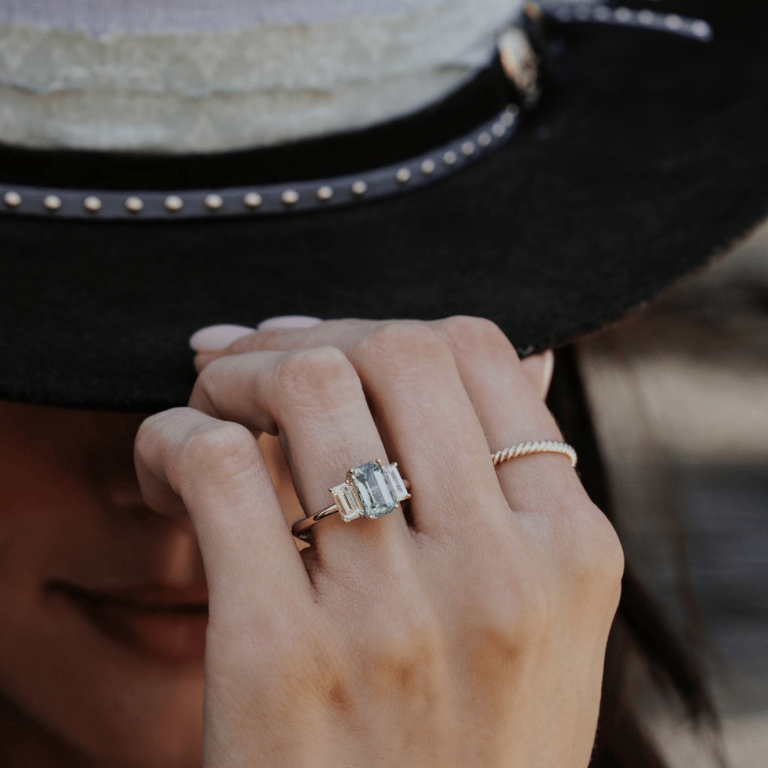 A 14k gold ring featuring a blue /green sapphire gemstone displayed on the ring finger of a hand model. 