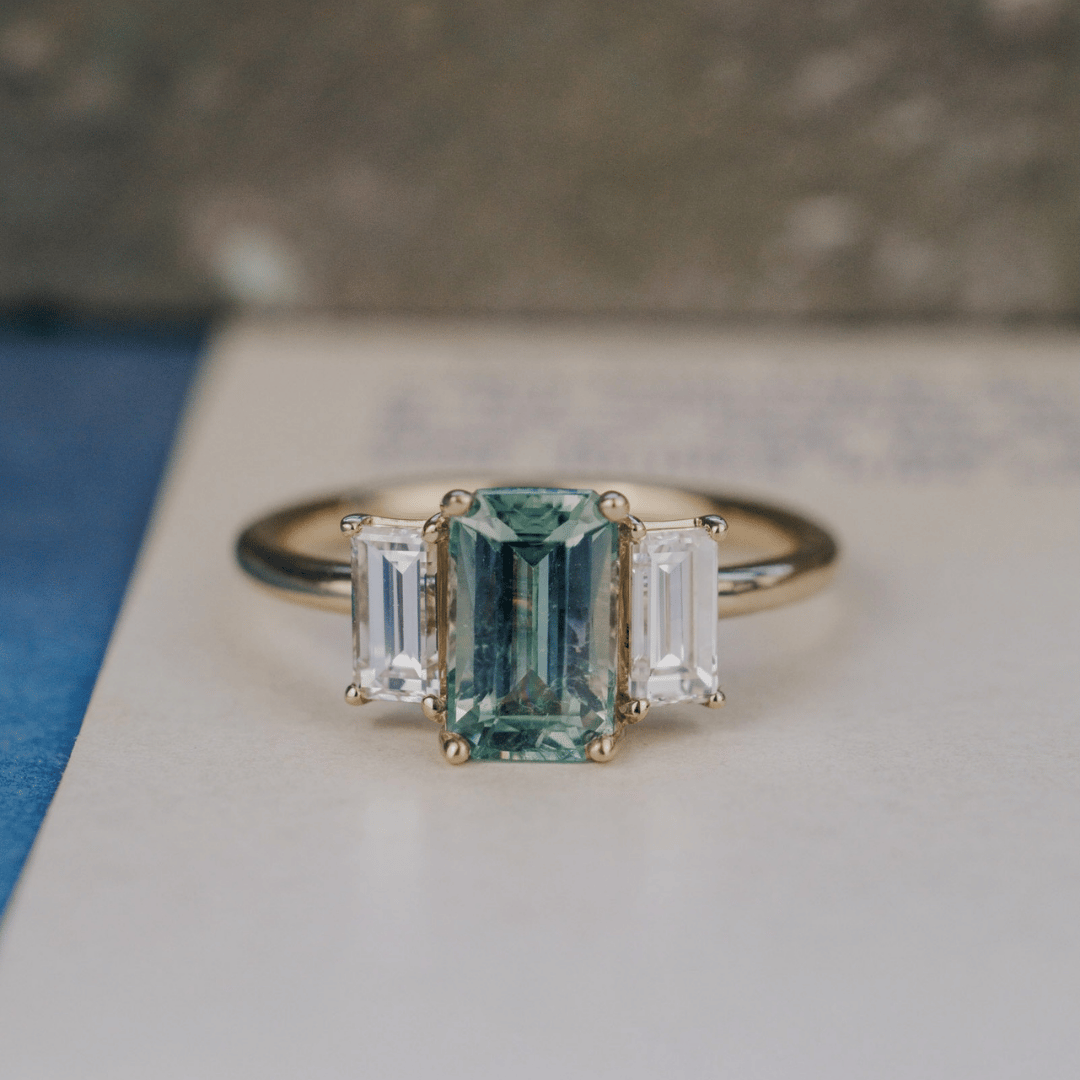 A 14k gold ring featuring a blue /green sapphire gemstone viewed from straight on, displayed on a textured white surface with a soft-focus background.