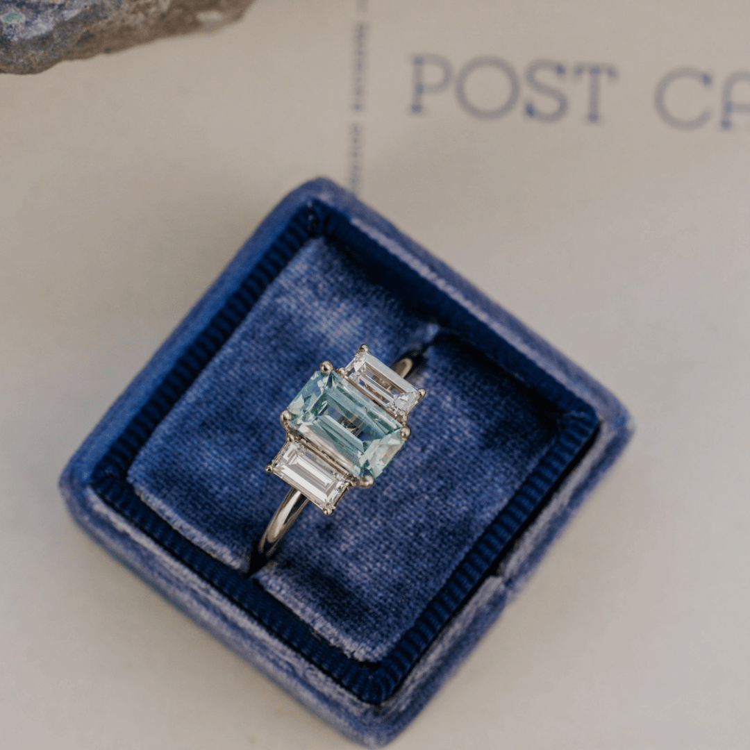 A 14k gold ring featuring a blue /green sapphire gemstone viewed from a ring box, displayed on a textured white surface with a soft-focus background.