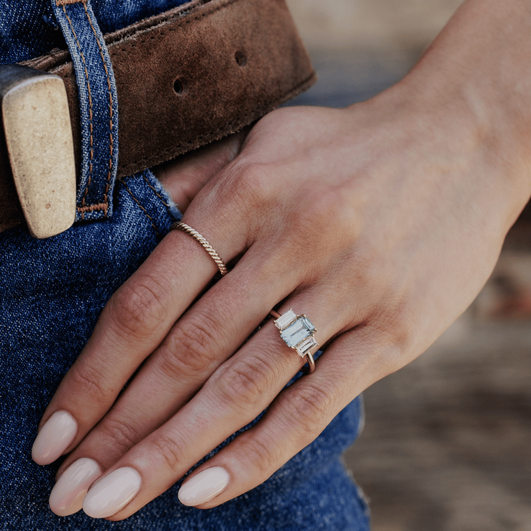 A 14k gold ring featuring a blue/green sapphire gemstone featured on the ring finger of a hand model. 