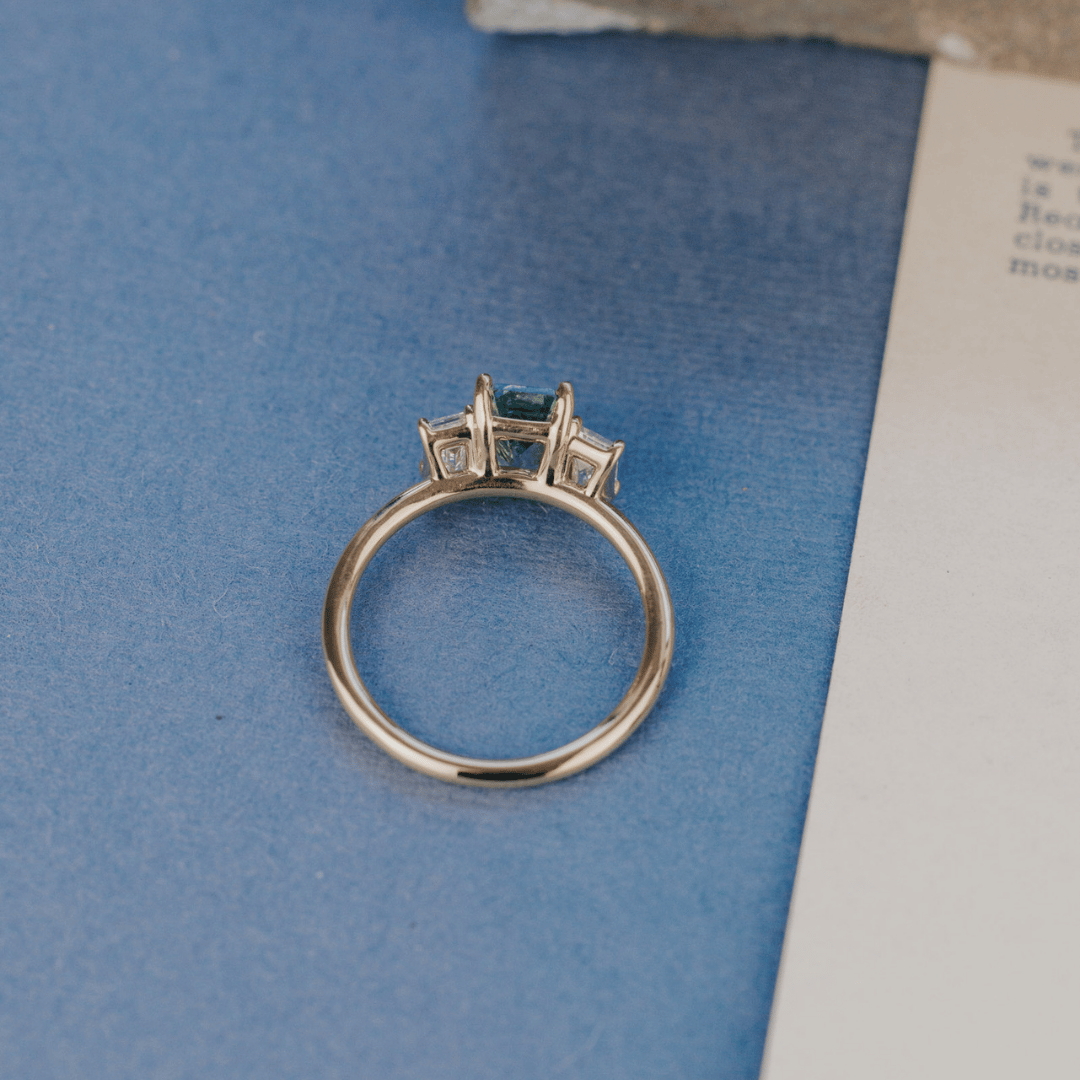 A 14k gold ring featuring a blue /green sapphire gemstone viewed from the top, displayed on a textured blue and white surface with a soft-focus background.
