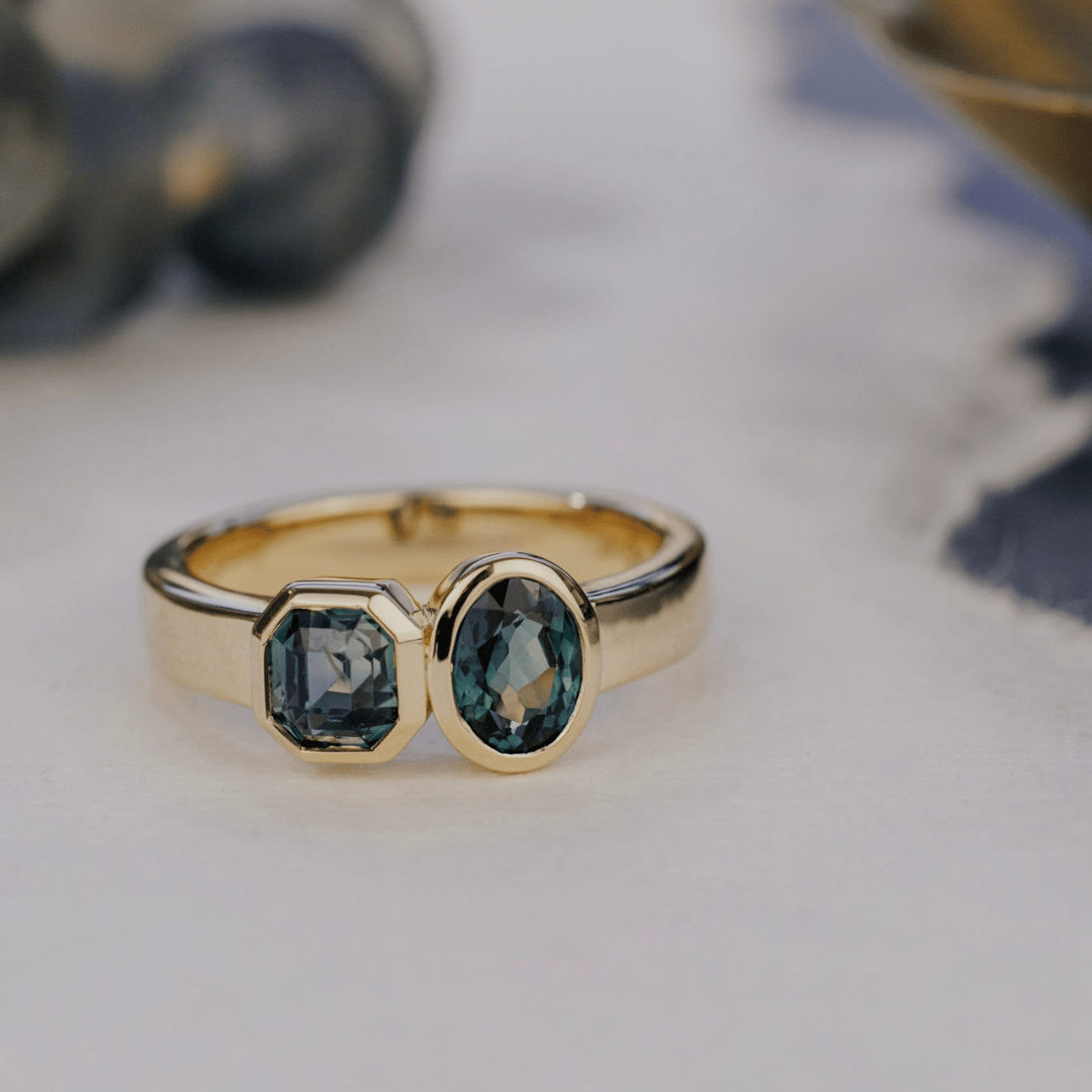 A 14k gold, two-stone ring with bezel-set Asscher and oval blue/teal sapphires in yellow gold. Set on a white surface with a soft-focus background. 