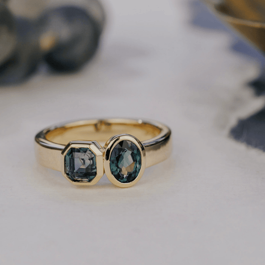 A 14k gold, two-stone ring with bezel-set Asscher and oval blue/teal sapphires in yellow gold. Set on a white surface with a soft-focus background. 
