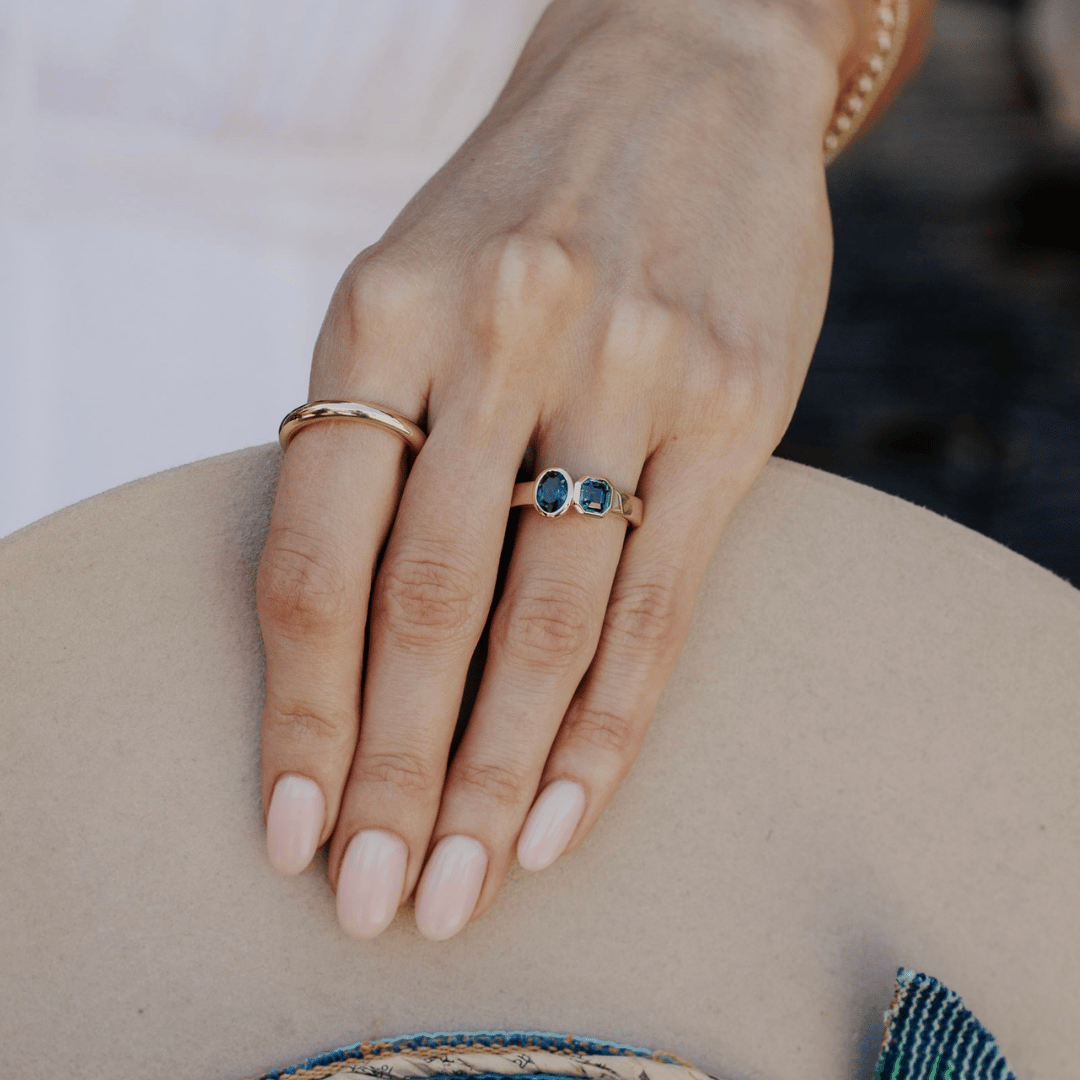 A 14k gold, two-stone ring with bezel-set Asscher and oval blue/teal sapphires in yellow gold. Set on the ring finger of a hand model, holding a hat.  