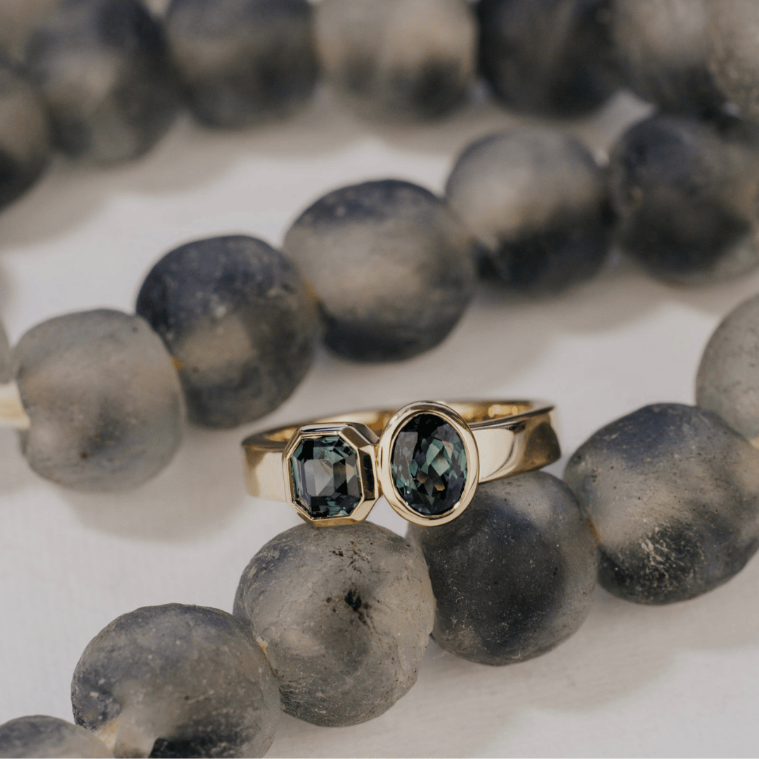 A 14k gold, two-stone ring with bezel-set Asscher and oval blue/teal sapphires in yellow gold. Set on a white surface on dark beads with a soft-focus background. 