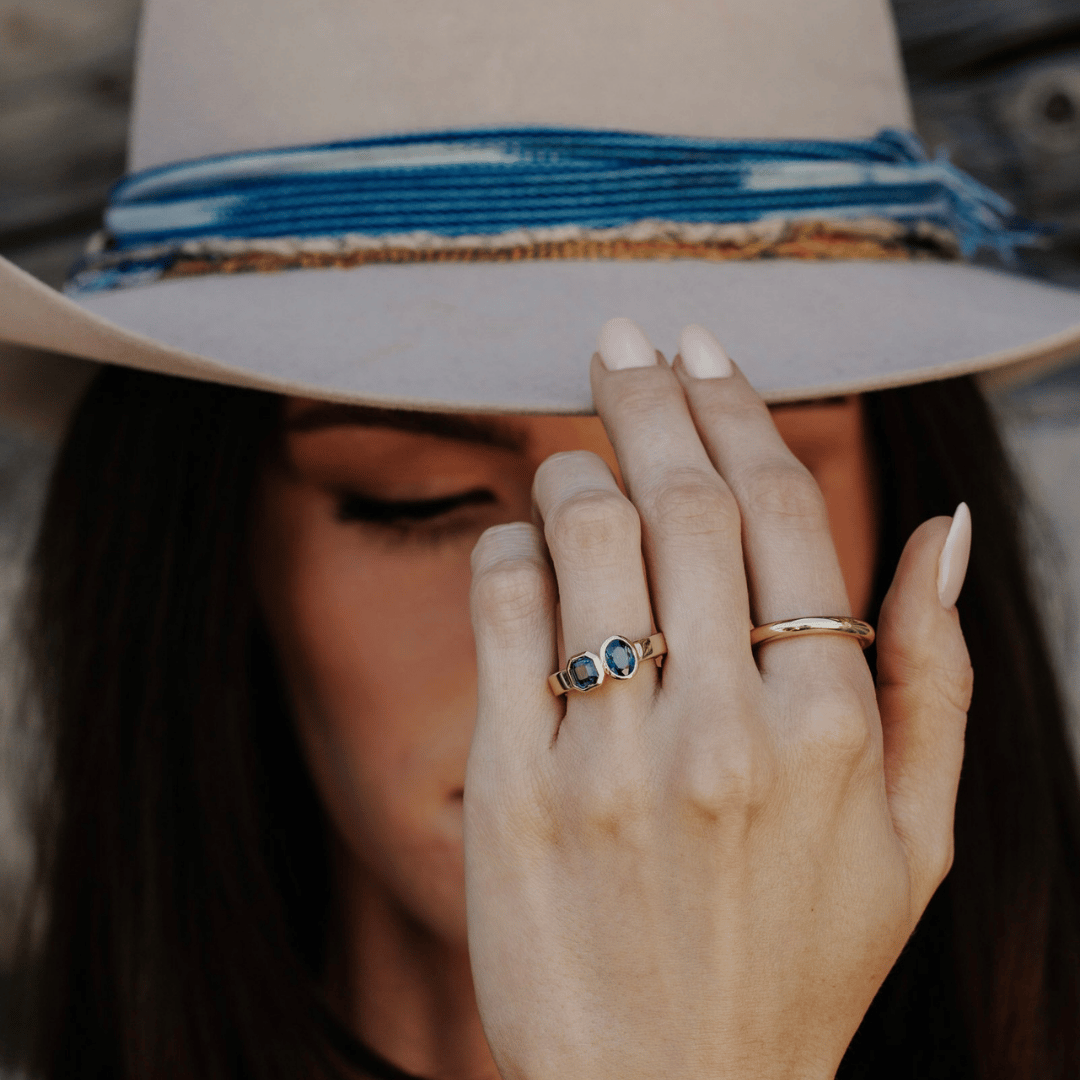A 14k gold, two-stone ring with bezel-set Asscher and oval blue/teal sapphires in yellow gold. Shown on the ring finger of a hand model, tipping the brim of her hat. 