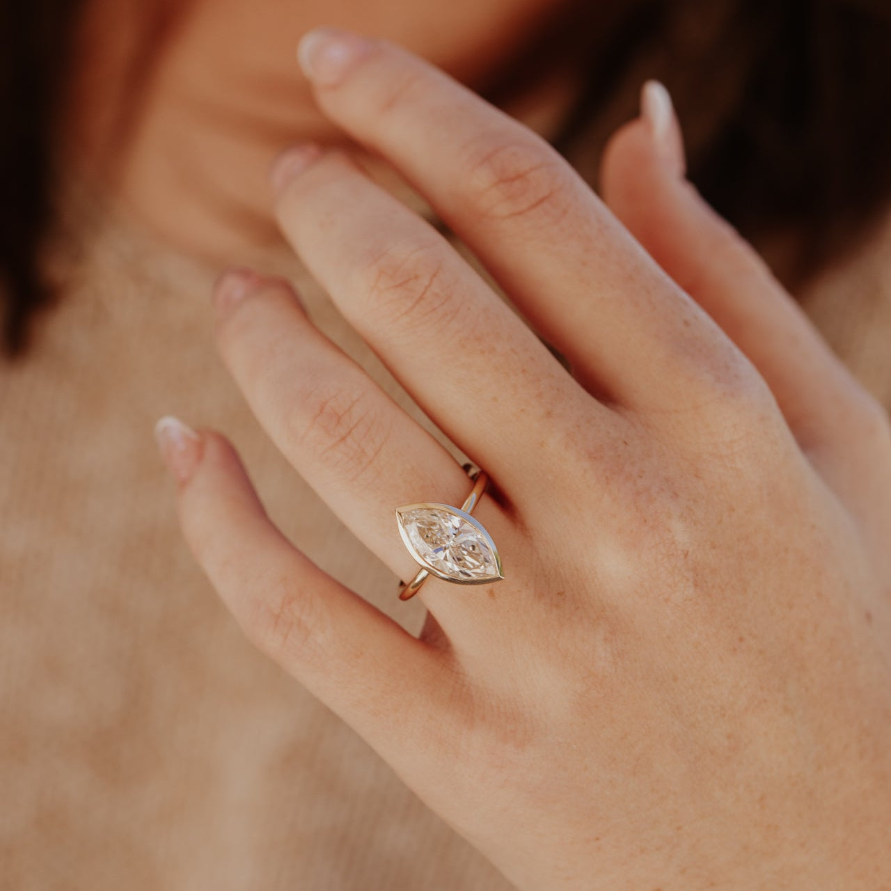 Close-up of a hand wearing a gold ring with a marquise diamond on a blurred background