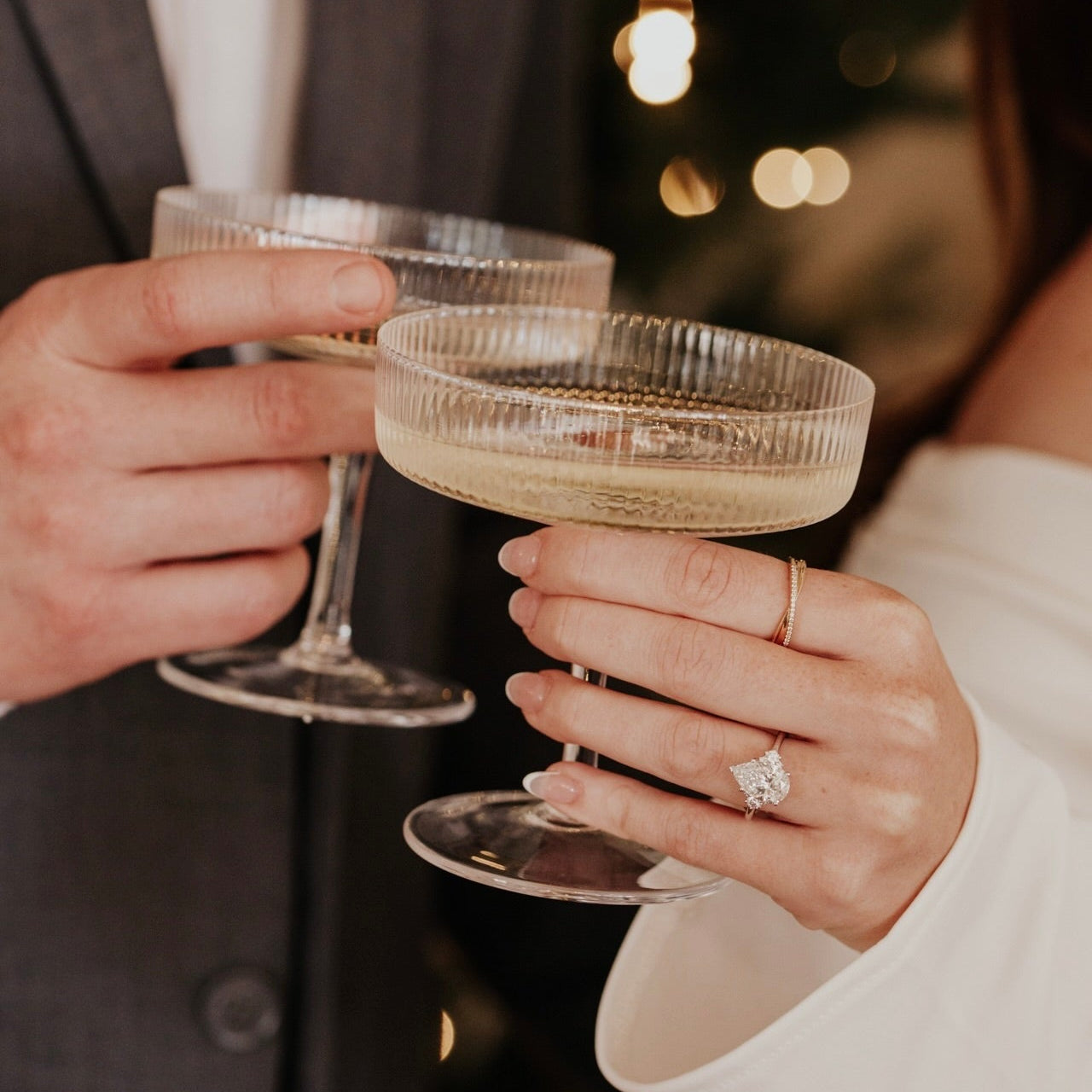 Two people clinking champagne glasses with a blurred festive background wearing a pear shaped lab grown diamond ring