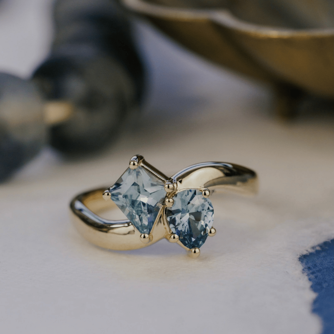A 14k gold ring featuring kite-shaped and pear-shaped blue sapphire gemstones, displayed on a texteured white background with a soft background. 