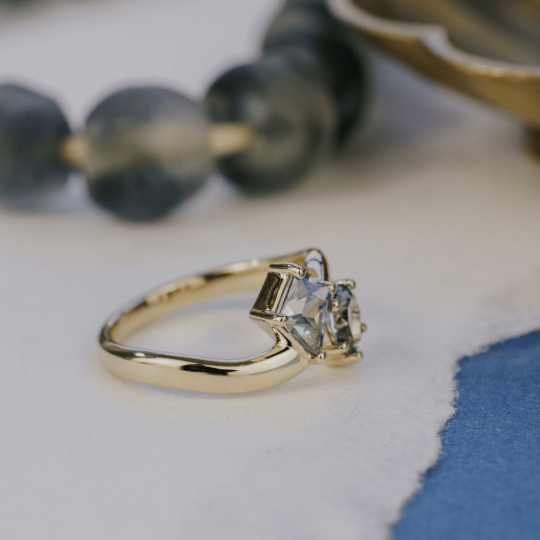 A 14k gold ring featuring kite-shaped and pear-shaped blue sapphire gemstones, turned at an angle, displayed on a texteured white background with a soft background. 