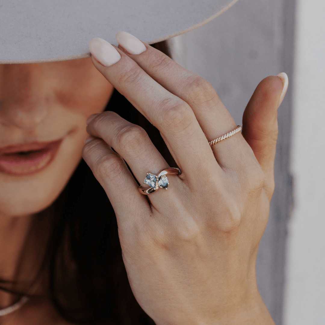 A 14k gold ring featuring kite-shaped and pear-shaped blue sapphire gemstones, displayed on the ring finger of a hand model with a soft-focus background.