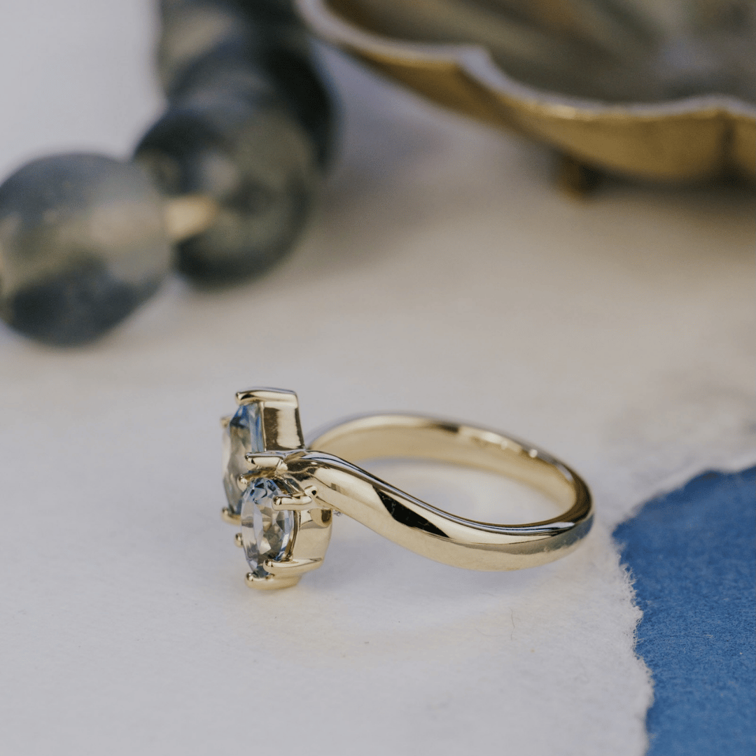 A 14k gold ring featuring kite-shaped and pear-shaped blue sapphire gemstones, turned to the side profile, displayed on a texteured white background with a soft background. 