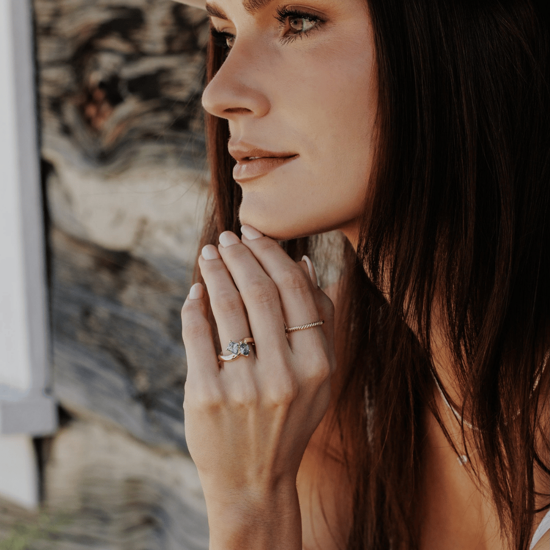A 14k gold ring featuring kite-shaped and pear-shaped blue sapphire gemstones, displayed on the ring finger of a hand model, side profile, touching the tip of her chin.