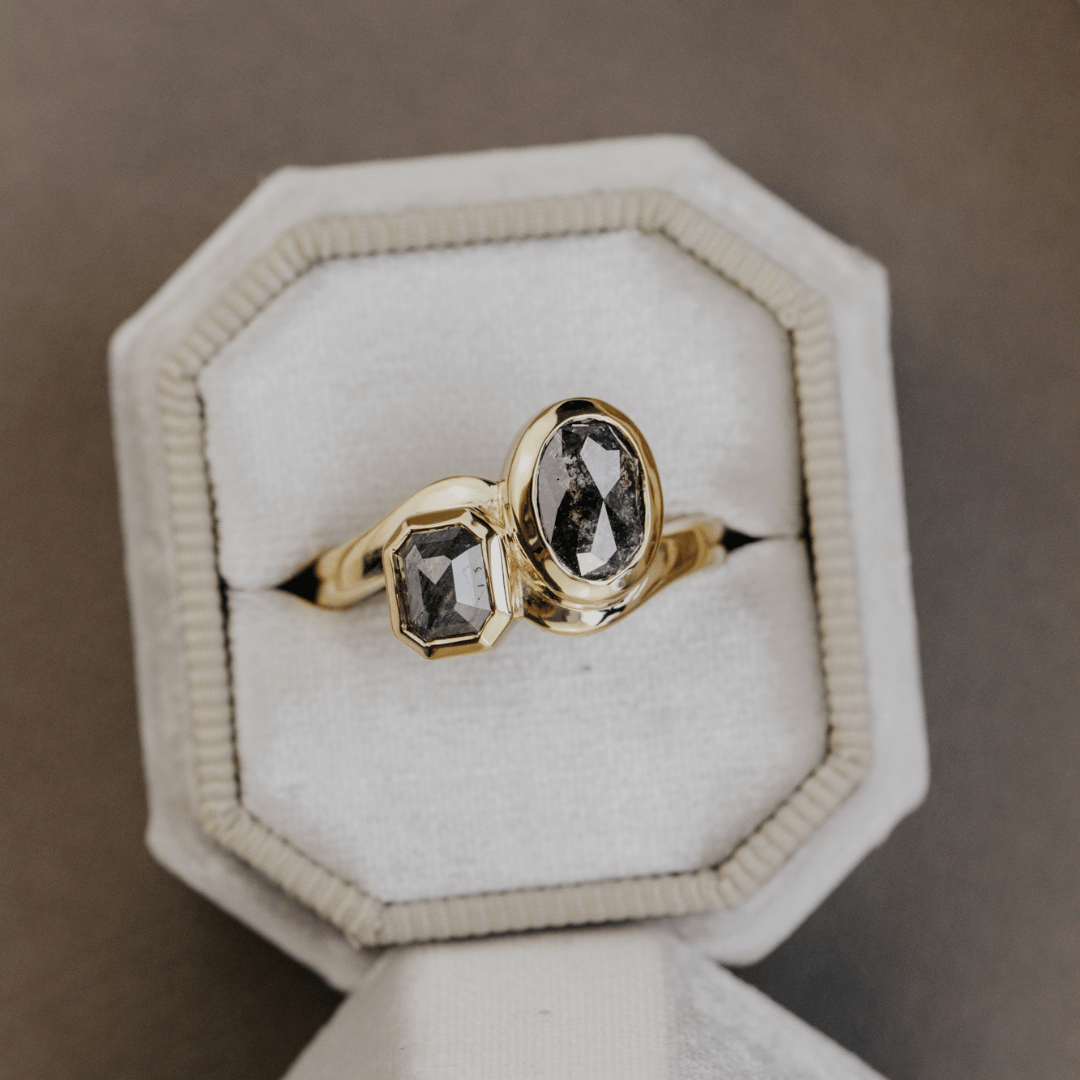 A gold ring with two dark gray rustic diamond gemstones, one oval and one rectangular, displayed in an octagonal cream-colored ring box.