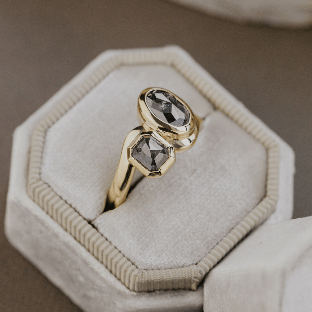 A gold ring with two faceted dark gray rustic diamonds displayed in a cream-colored jewelry box.