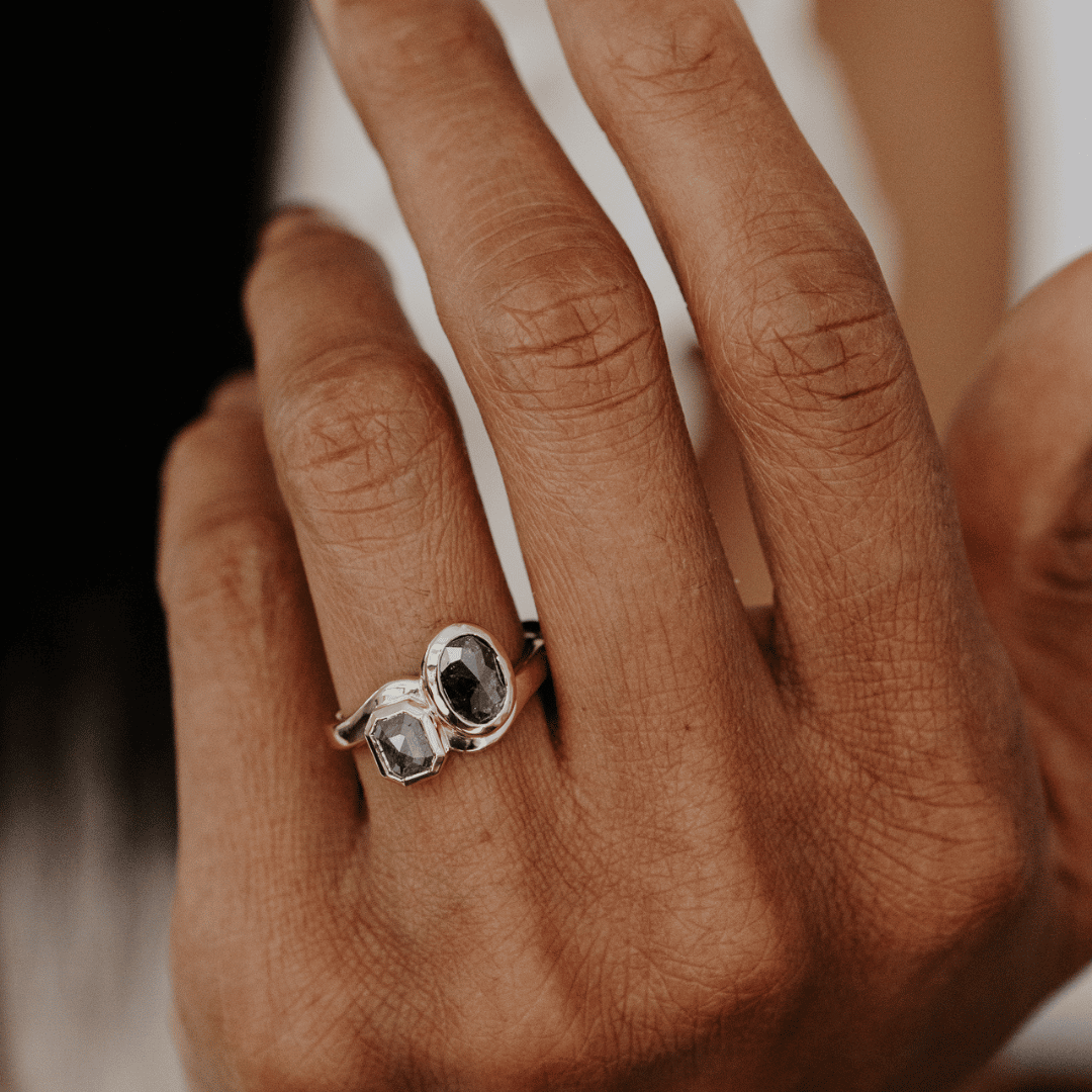 A close-up of a hand wearing a gold ring with two rustic diamonds, set against a softly blurred background.