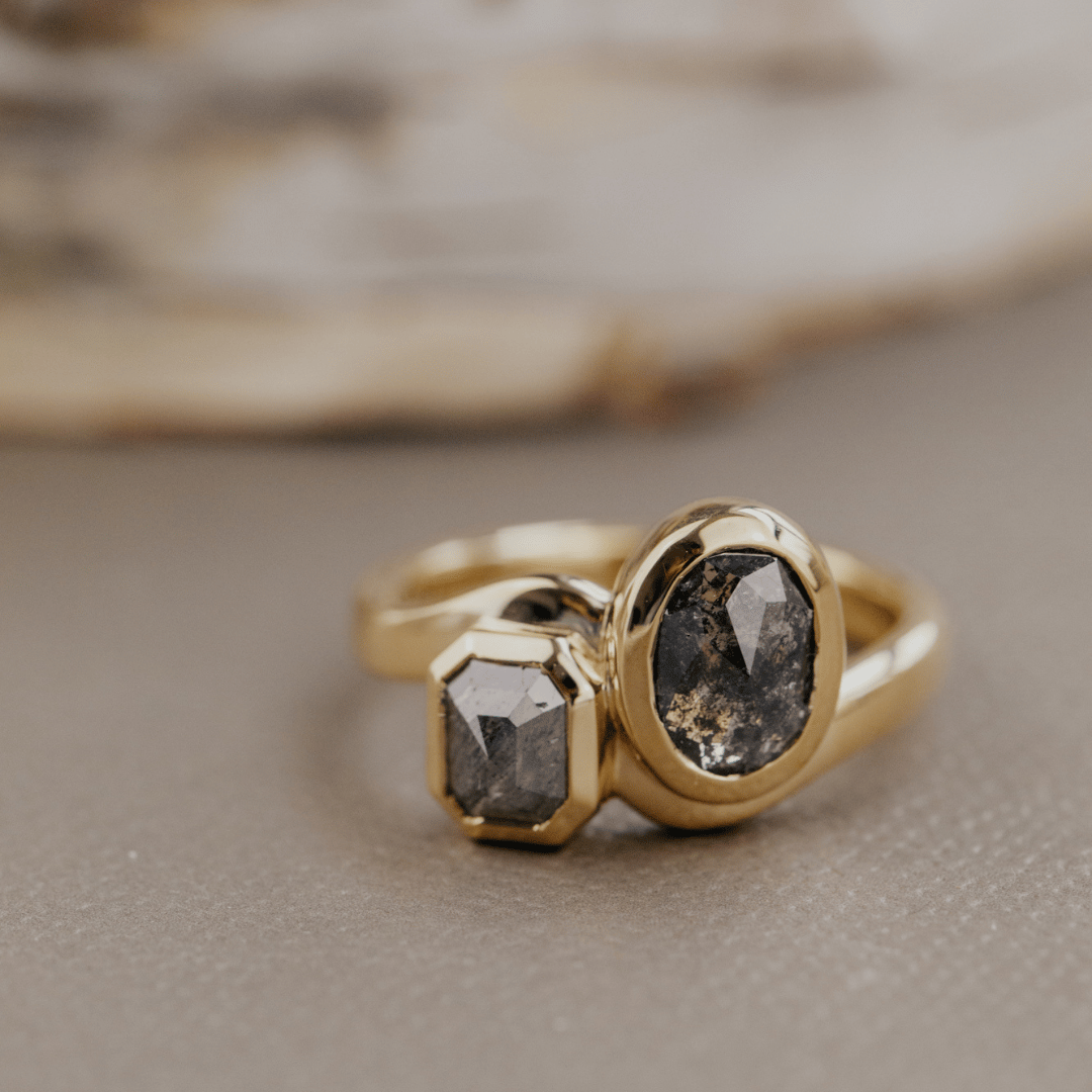 A gold ring with two dark gray gemstones, one oval and one rectangular, resting on a textured gray surface with a blurred natural background.