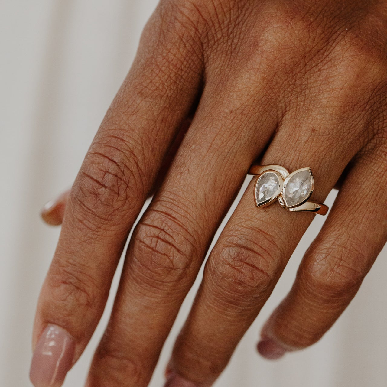 Close-up of a hand wearing a gold ring with two large rustic diamond gemstones on a neutral background.