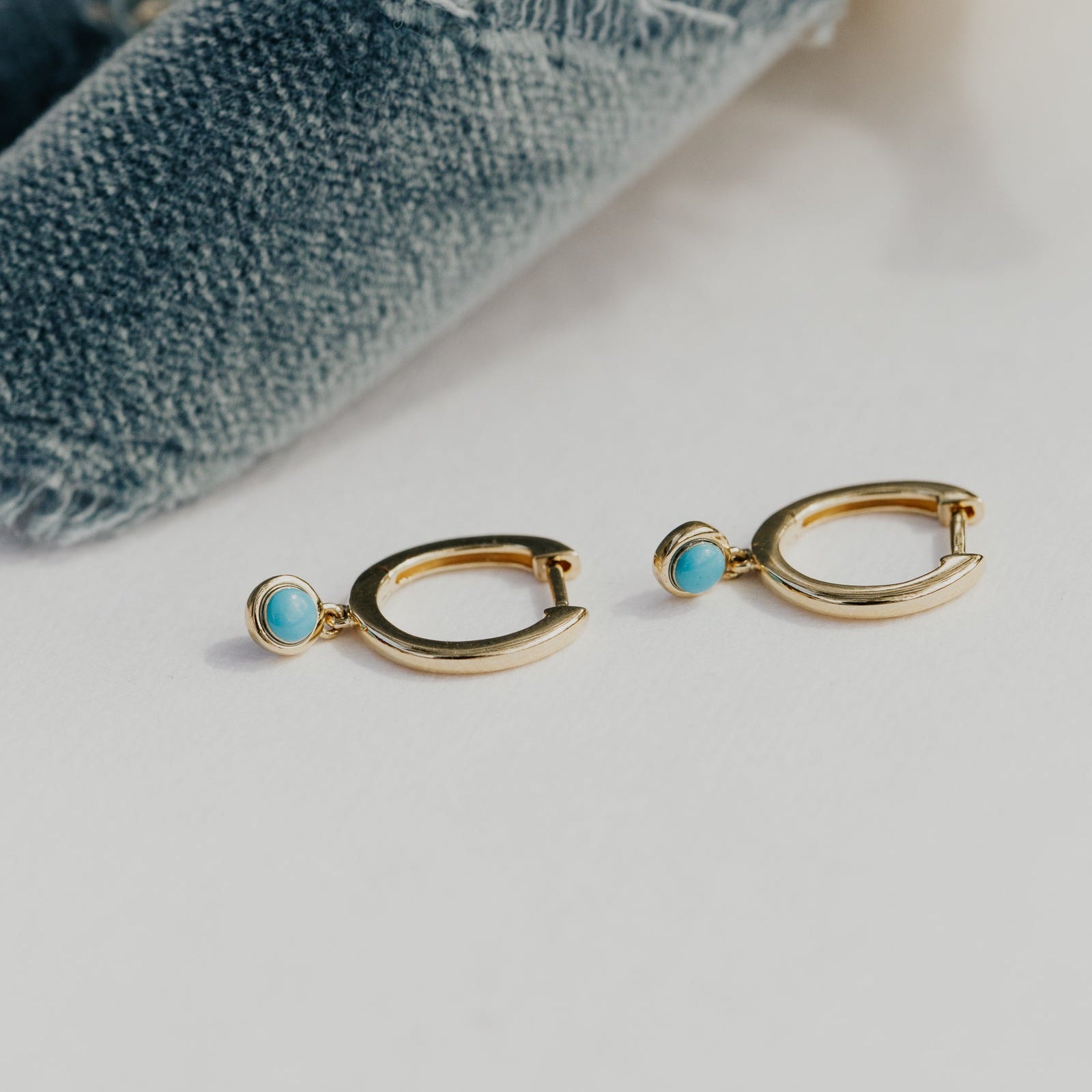 Gold hoop earrings with turquoise stones on a light surface with denim fabric in the background.