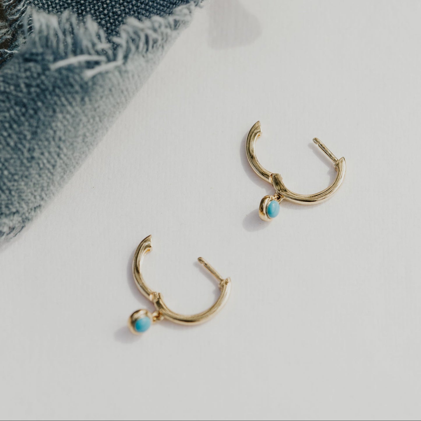 Gold hoop earrings with turquoise stones on a white surface with denim fabric.