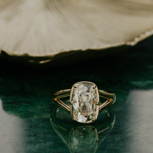 Gold ring with a elongated cushion old mine cut antique diamond on a green surface with a leaf in the background.