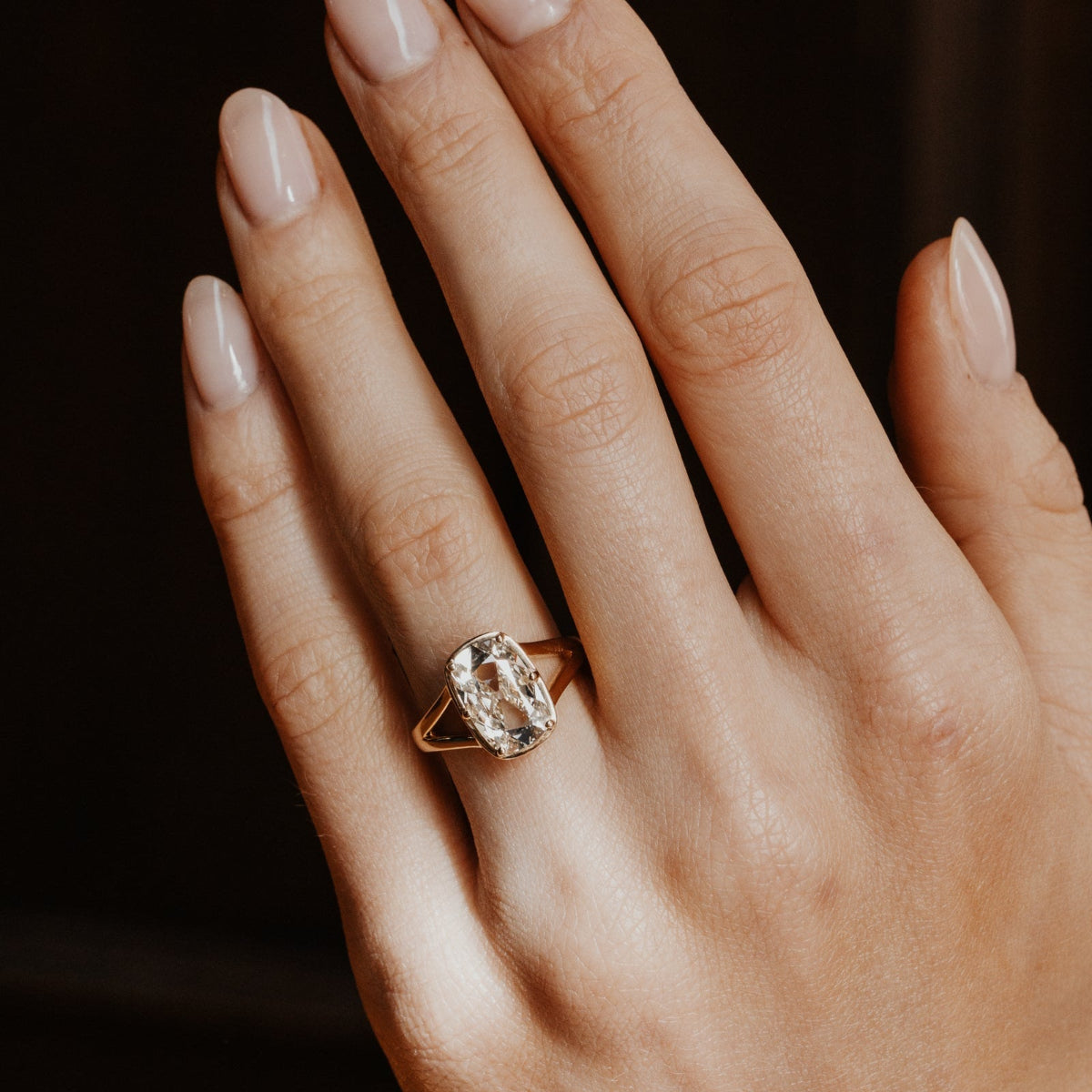 Hand wearing a gold ring with a elongated cushion old mine cut antique diamond on a dark background.