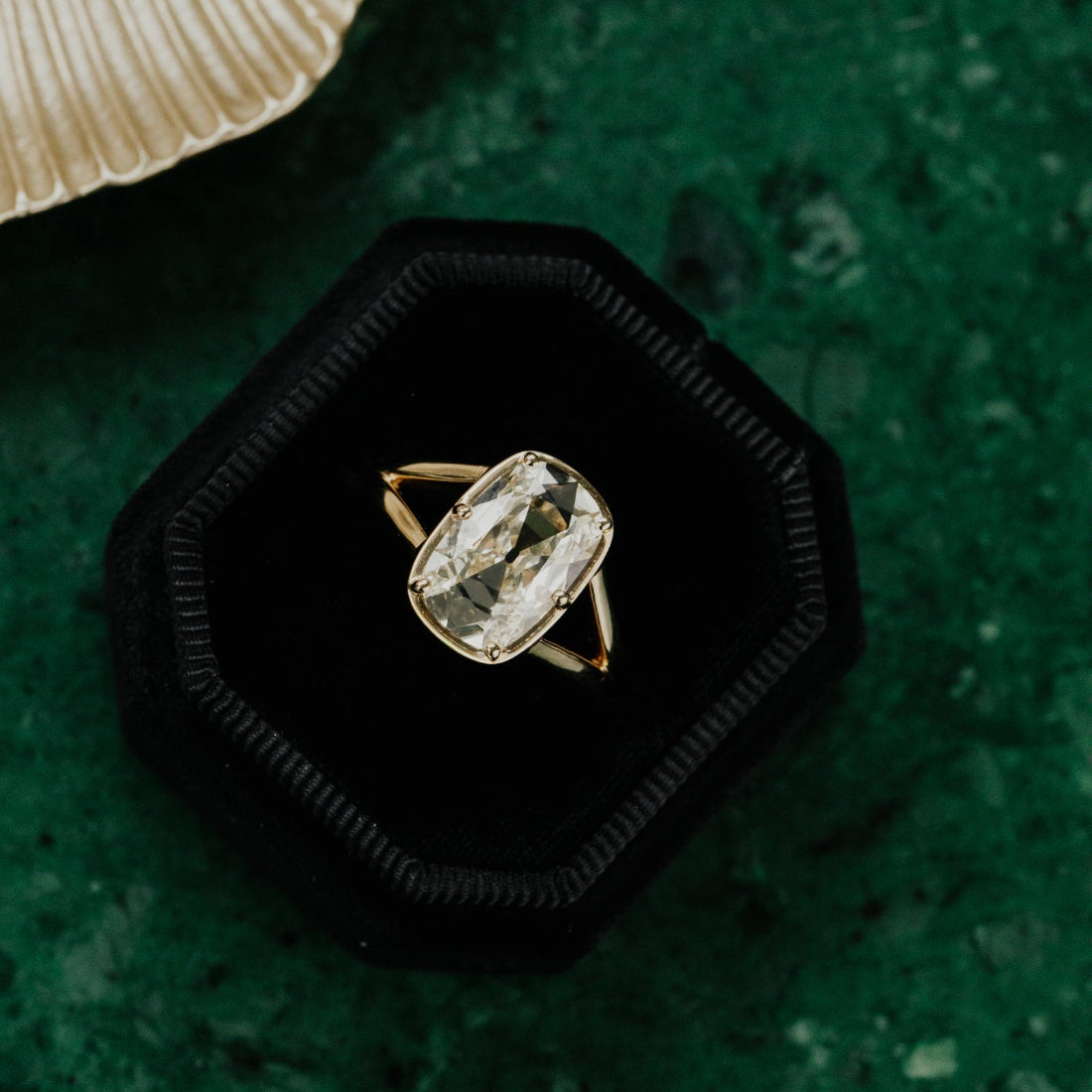 Gold ring with a elongated old mine cut antique diamond in a black hexagonal box on a green textured surface.