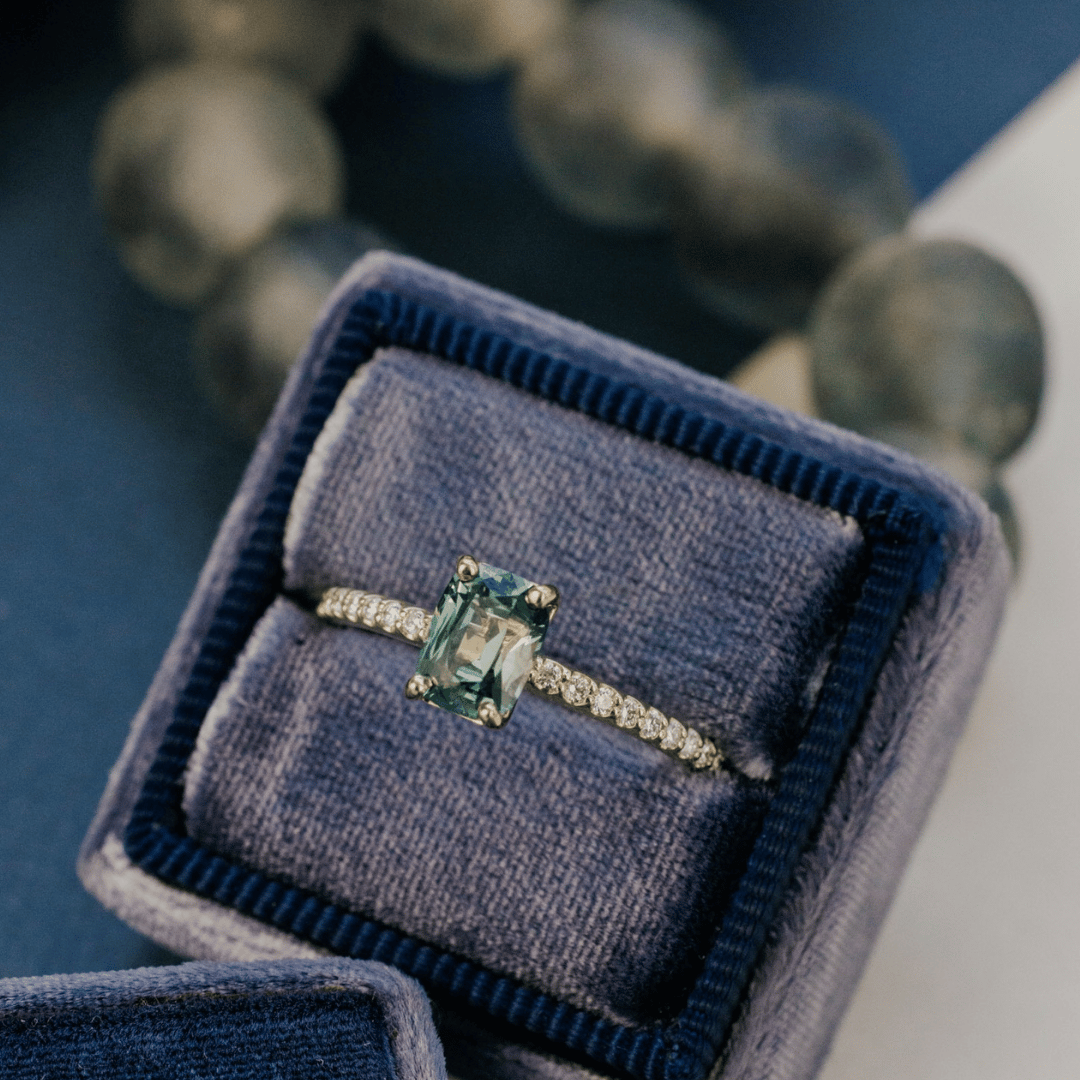 A 14k gold ring featuring a single blue sapphire gemstone from a top view, displayed in a ring box with a soft-focus background.