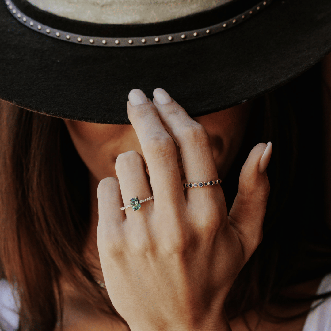 A 14k gold ring featuring a single blue sapphire gemstone, displayed on the ring finger of a hand model tipping her hat.