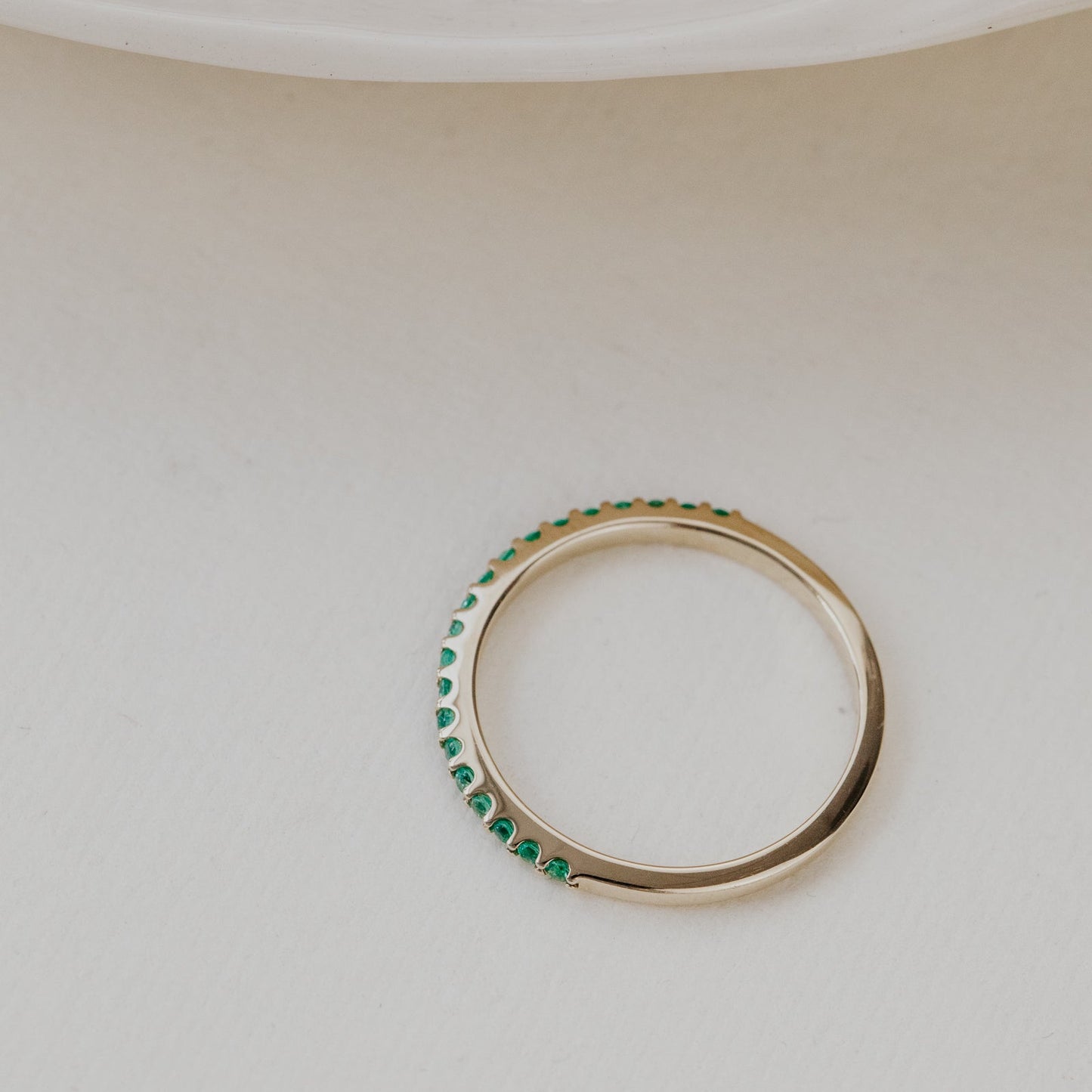 Gold wedding band with green emerald gemstones on a beige surface.