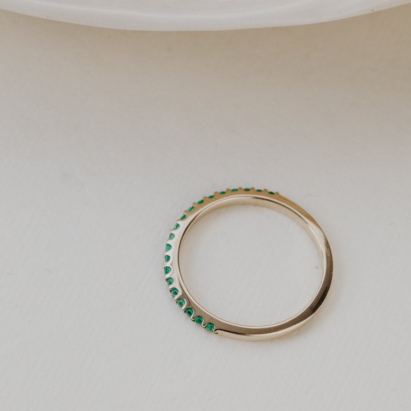 Gold wedding band with green emerald gemstones on a beige surface.