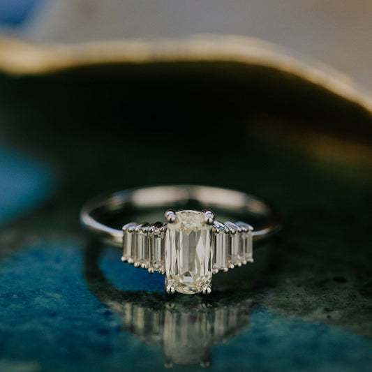 Diamond ring with a elongated cushion diamond center stone on a reflective surface.