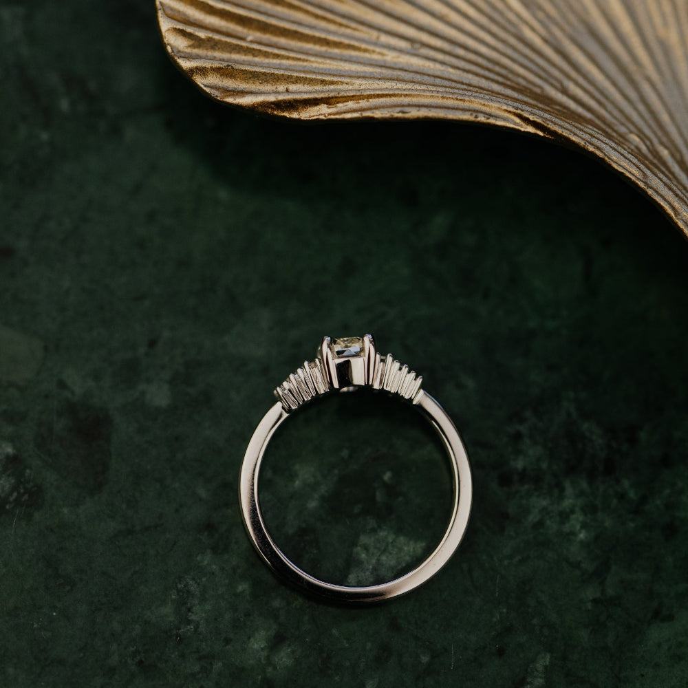 White gold ring with a diamond on a dark green surface with a ginkgo leaf.