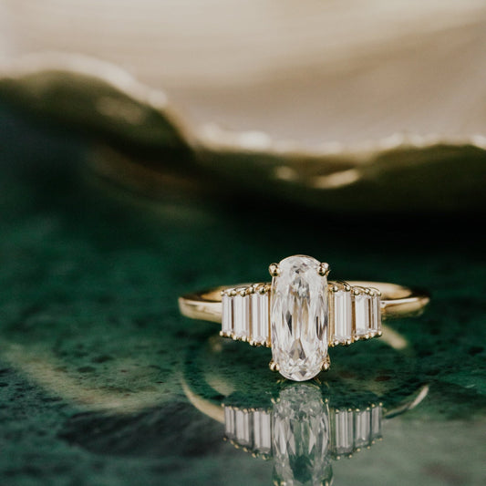 Gold Jolee ring with a Elongated cushion diamond on a green surface.