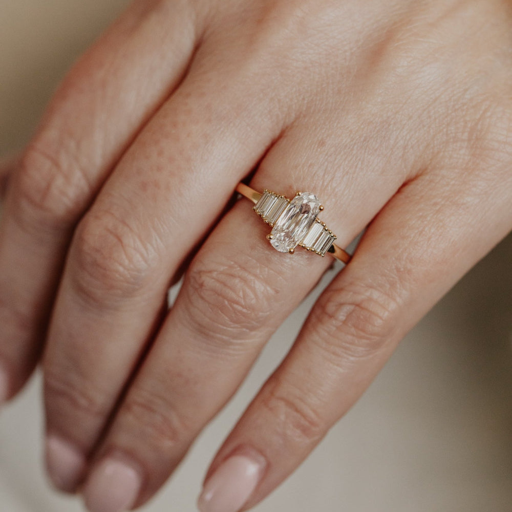 Close-up of a hand wearing a gold engagement ring with a elongated cushion diamond on a neutral background.