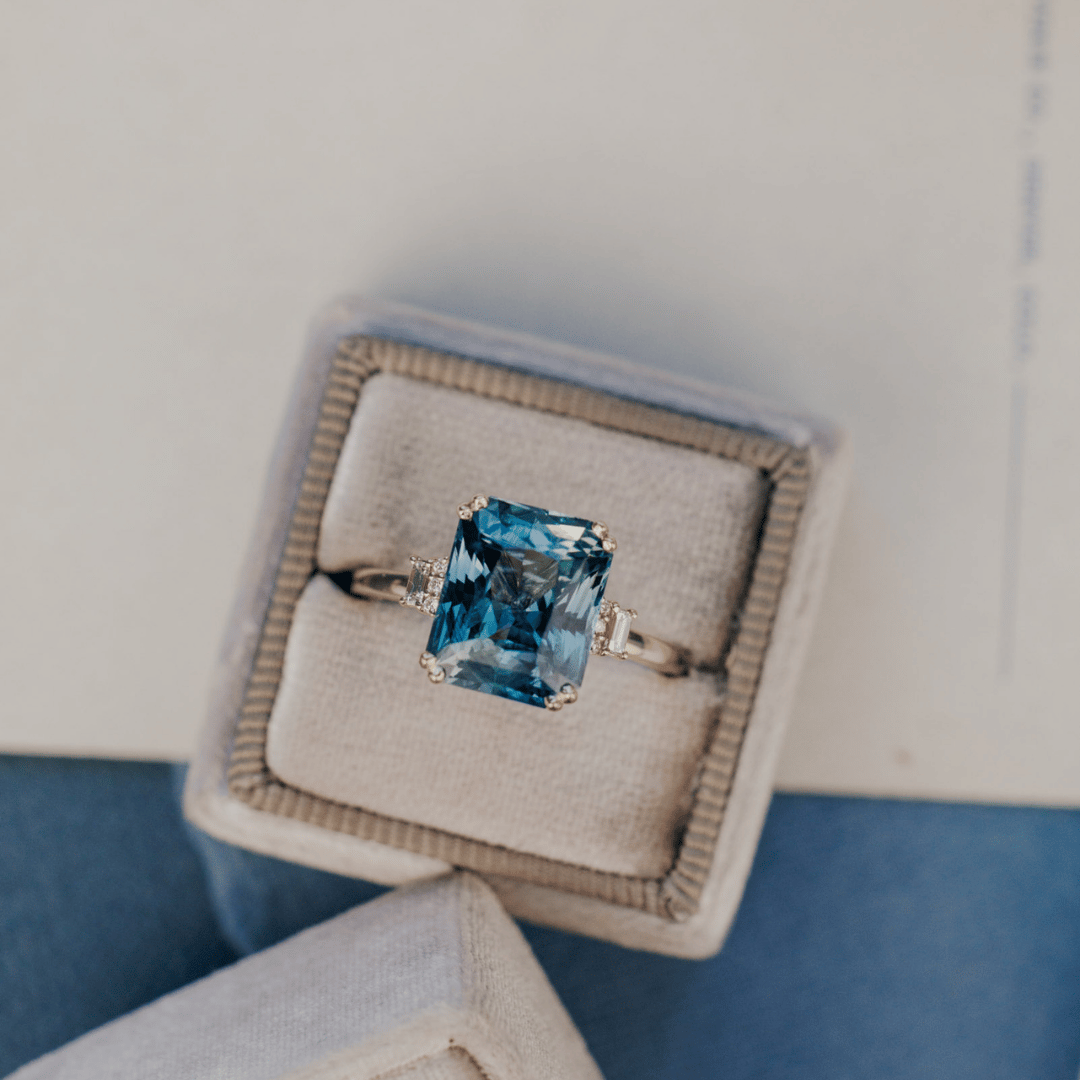 A 14k gold ring featuring a blue sapphire gemstone viewed from a ring box on a textured white surface.