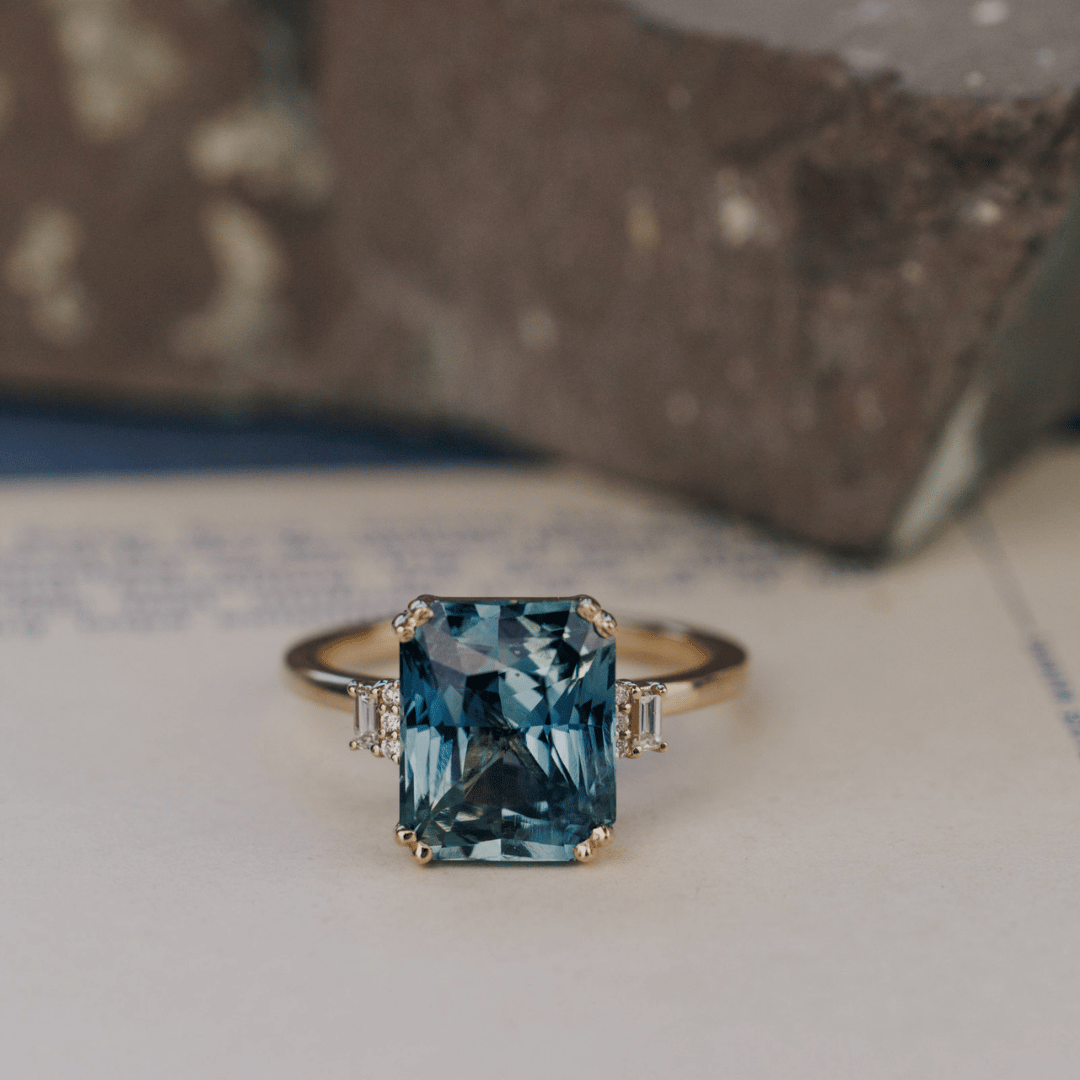 A 14k gold ring featuring a blue sapphire gemstone viewed from straight on, displayed on a textured white surface with a soft-focus background.