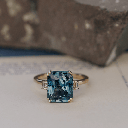 A 14k gold ring featuring a blue sapphire gemstone viewed from straight on, displayed on a textured white surface with a soft-focus background.