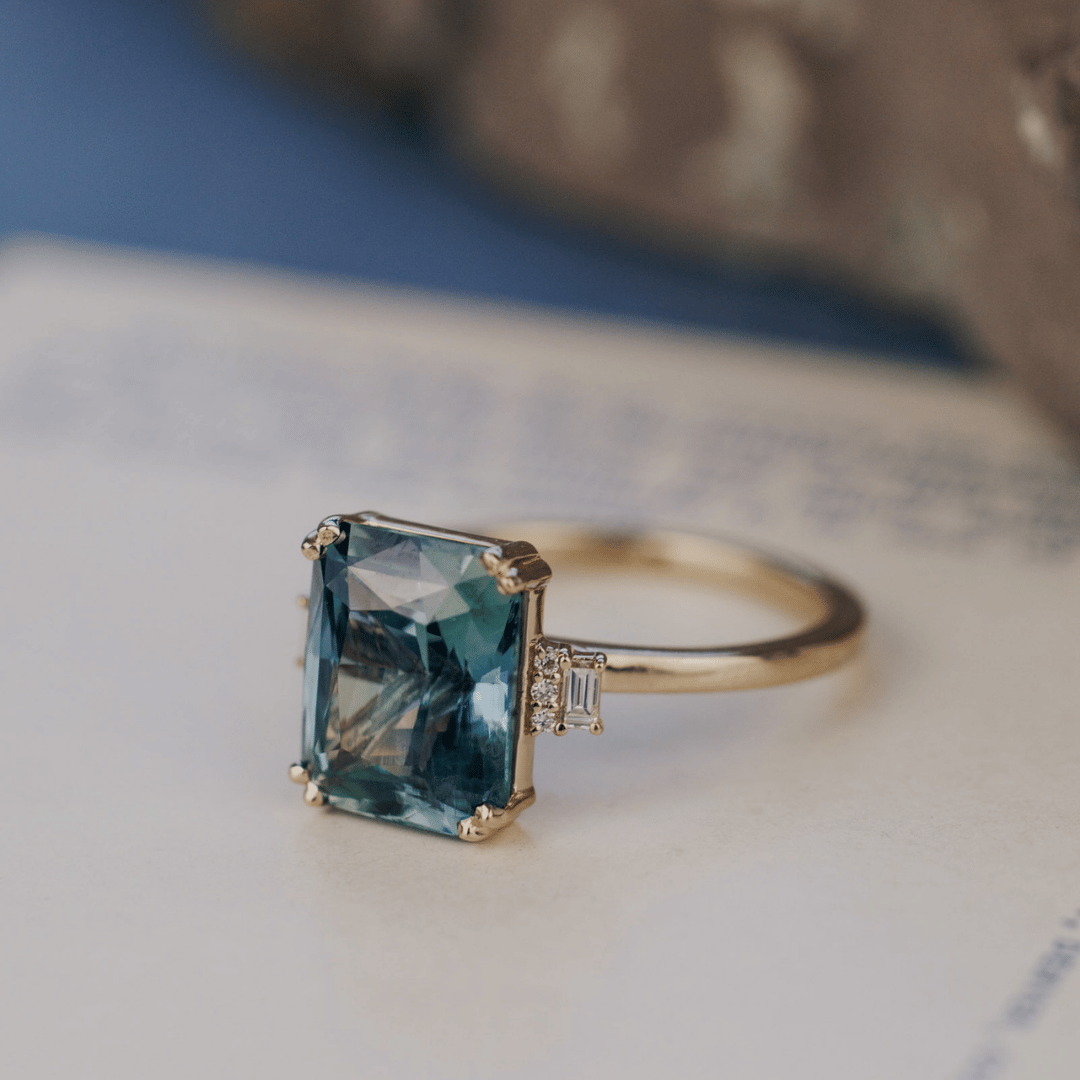 A 14k gold ring featuring a blue sapphire gemstone viewed from an angle, displayed on a textured white surface with a soft-focus background.