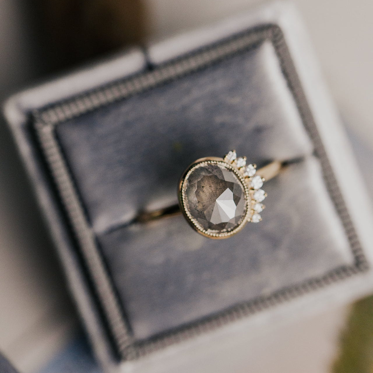Ring with a large oval gray rustic diamond with a diamond side flare on a gray velvet cushion.