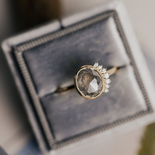 Ring with a large oval gray rustic diamond with a diamond side flare on a gray velvet cushion.