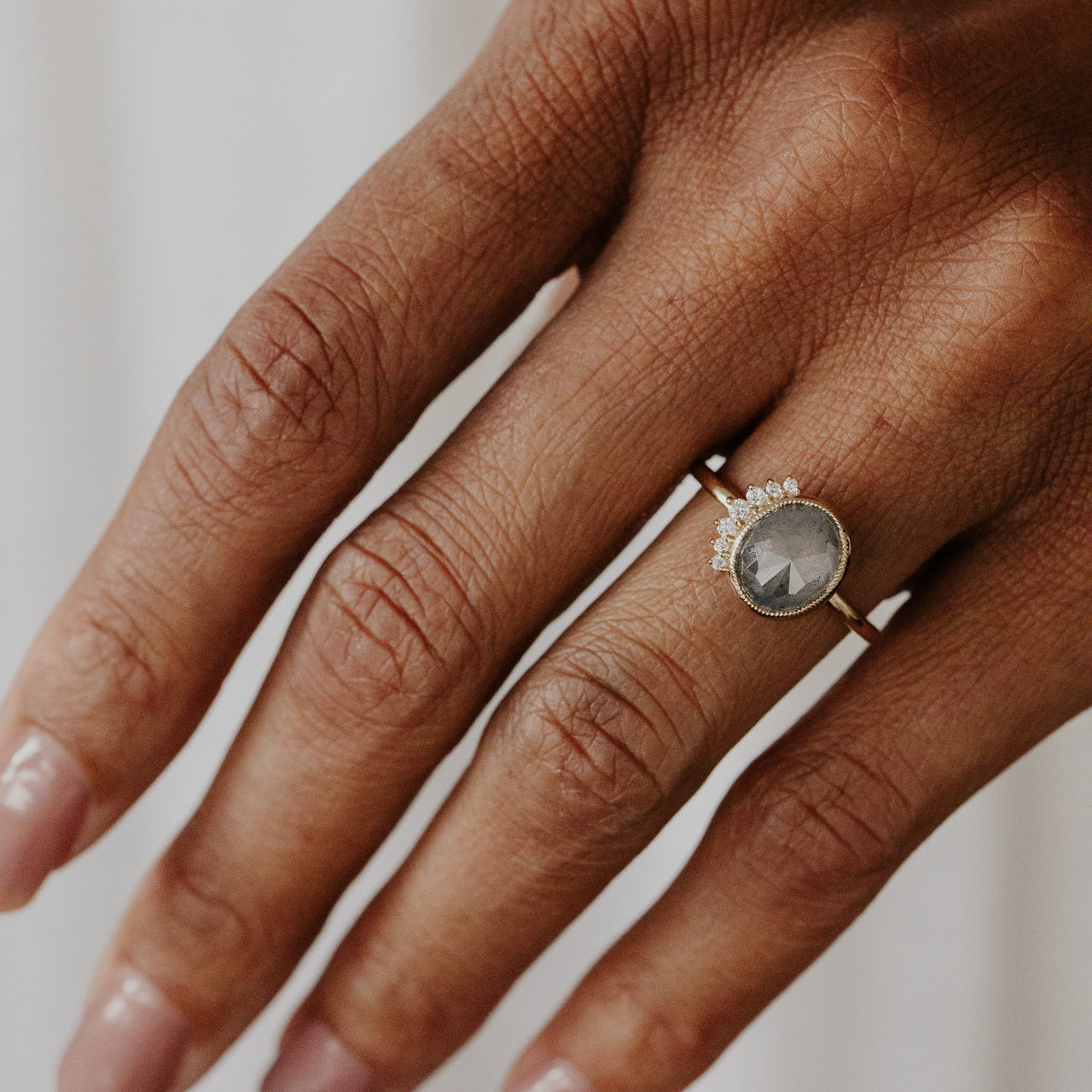 Hand wearing a ring with a oval gray rustic diamond on a neutral background.