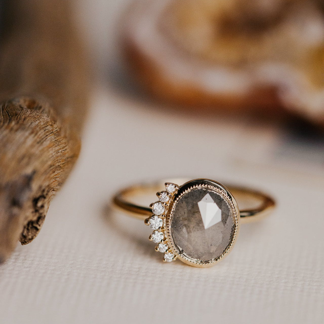Gold ring with a gray oval rustic diamond with a diamond side flare on a textured surface.