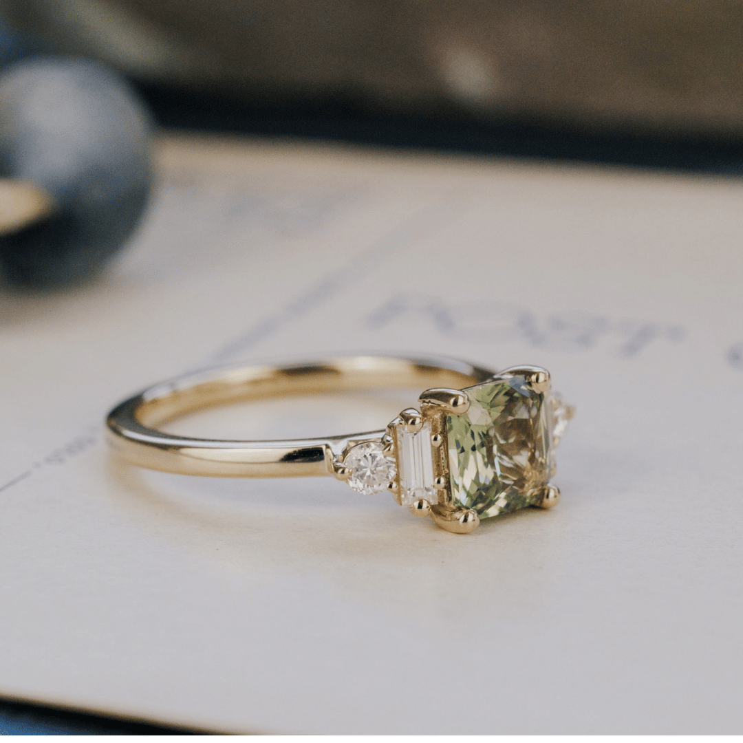 A 14k gold ring featuring a yellow-green sapphire gemstone, from the side view, displayed on a white surface with a soft background. 