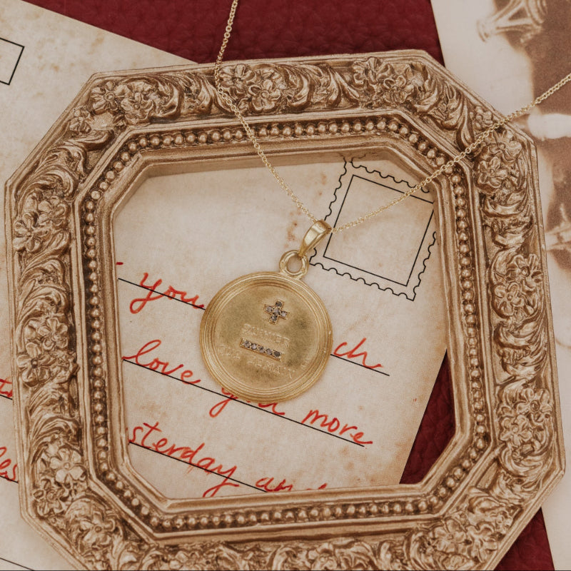 The Large 18k Yellow Gold Vintage Love Token with Diamonds by A. Augis, a round 18k yellow gold pendant with rose-cut natural diamonds and the engraved phrase "qu'hier que demain," styled on a gold chain atop a vintage love letter and antique photographs within an ornate gold frame.