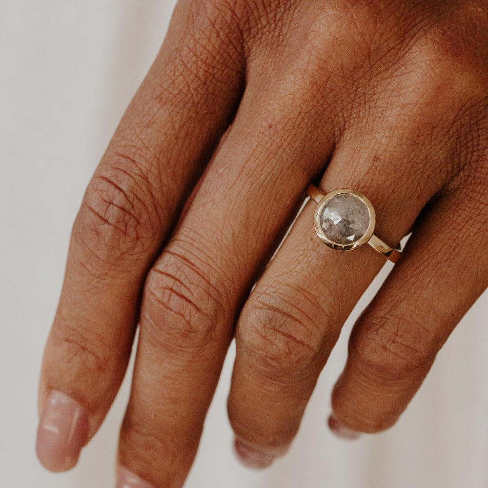 Hand wearing a gold ring with a round gray rustic diamond on a neutral background.