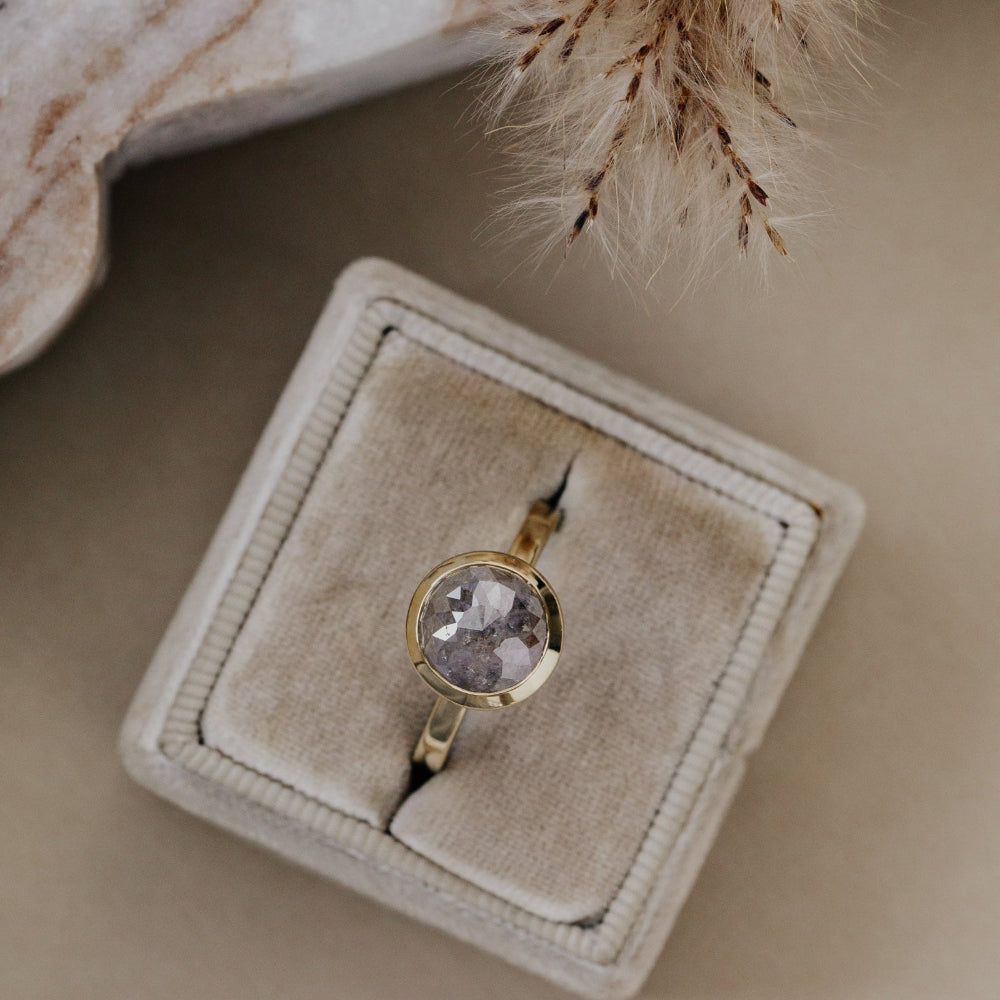 Gold ring with a round gray rustic diamond on a beige velvet cushion.