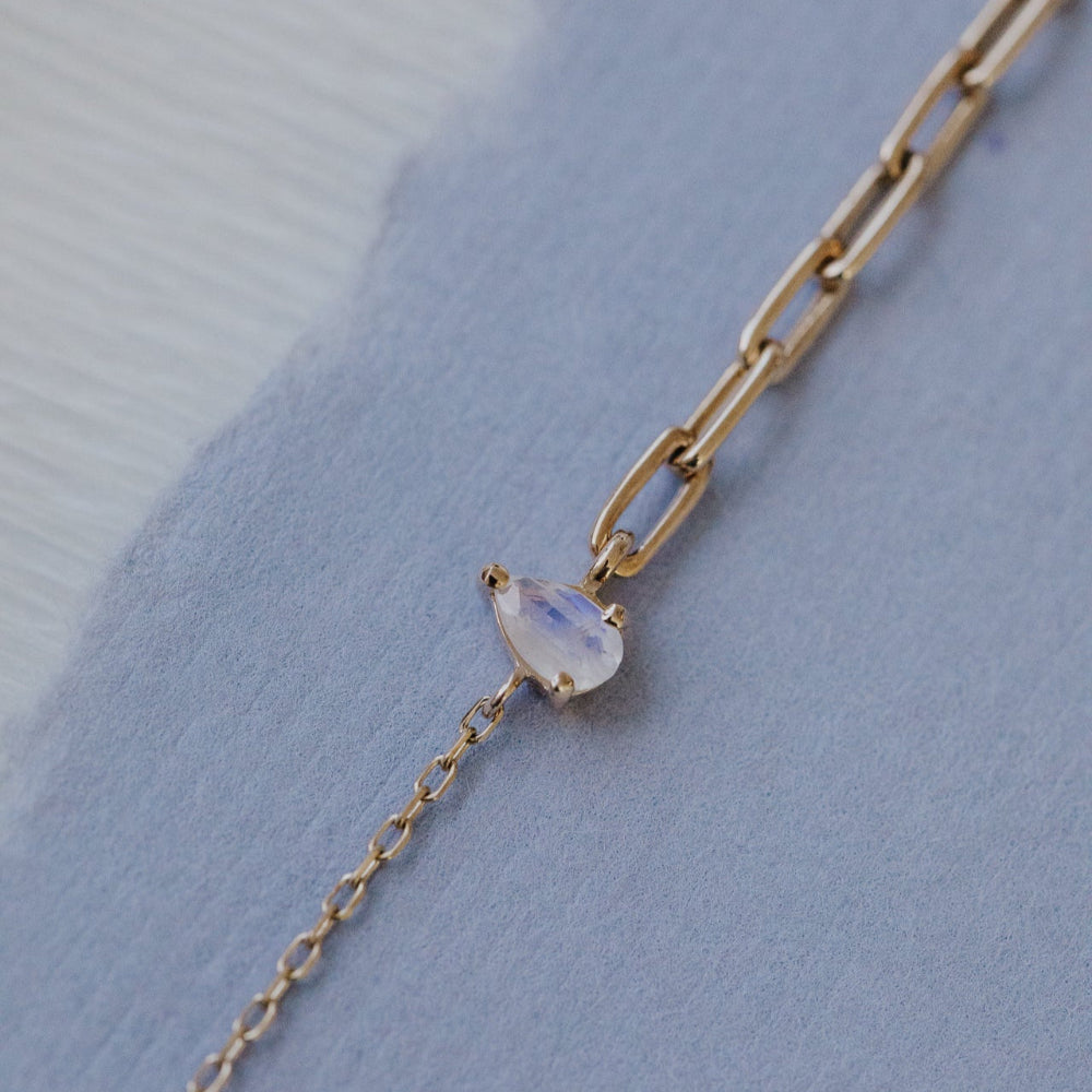 Gold chain necklace with a teardrop-shaped moonstone pendant on a light blue background